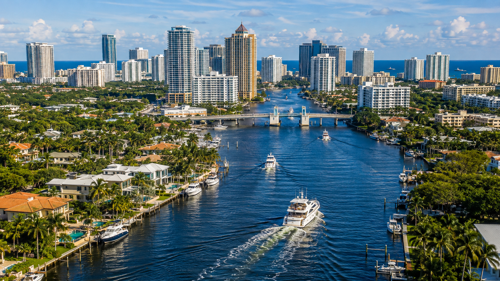 Aerial view of a coastal city with high-rise buildings lining a wide canal, dotted with boats and palm-lined homes nearby under a blue sky.
