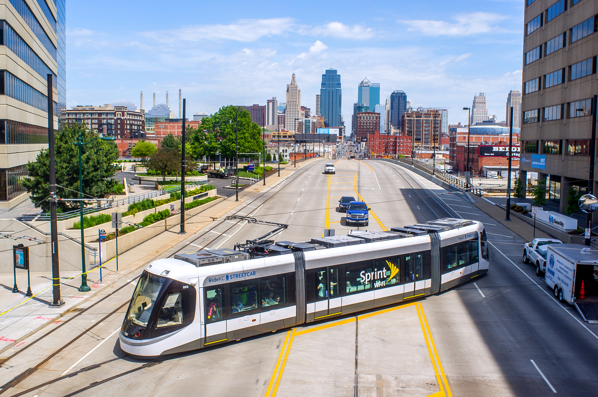 White sprint streetcar on tracks moves through a downtown avenue with a blue-sky skyline in the background beneath tall buildings and streetlamps: informative transport scene in city core.