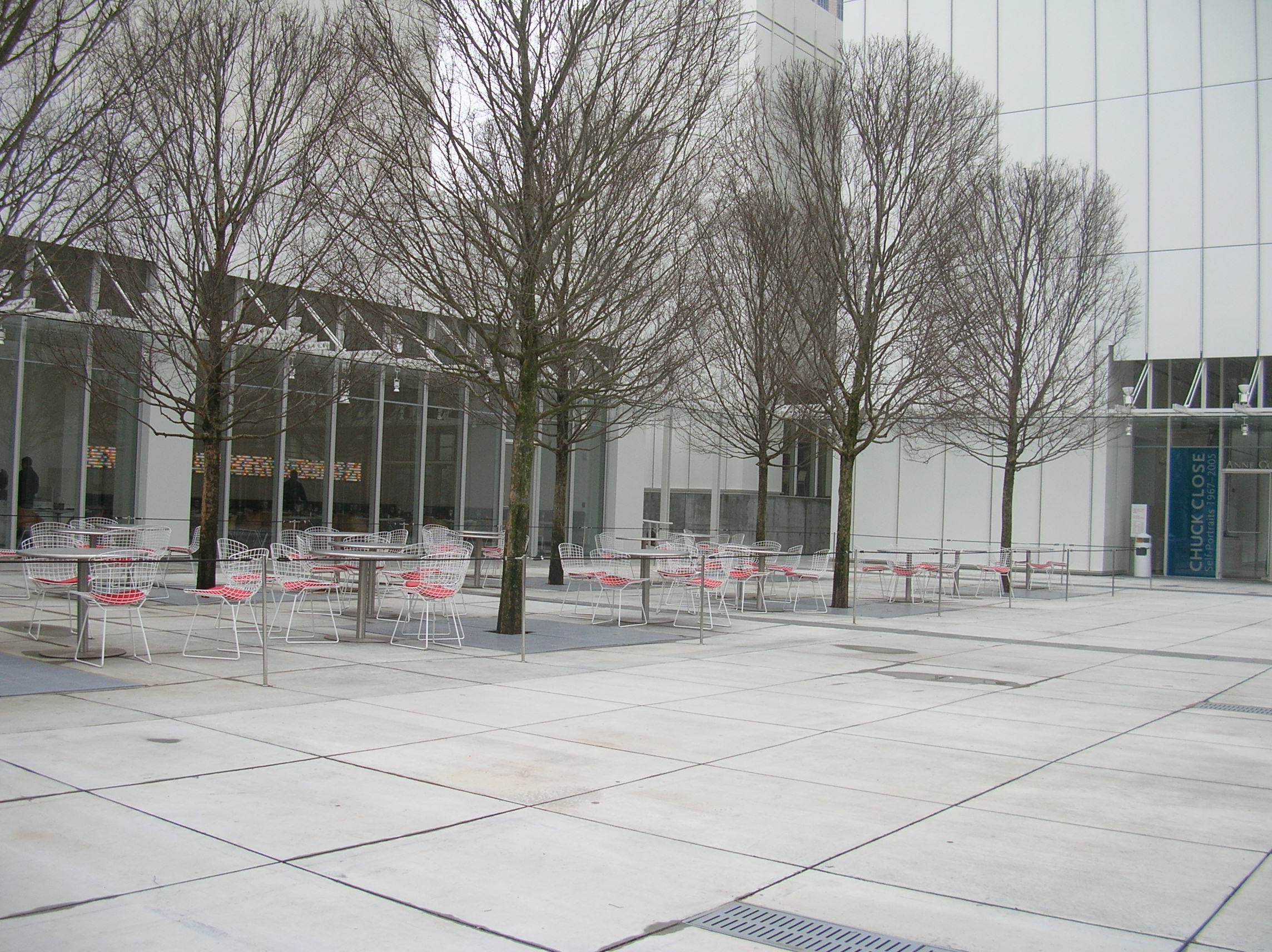 Outdoor cafe seating on a gray tiled plaza with white metal tables and chairs; bare trees line the area in front of a modern glass building.