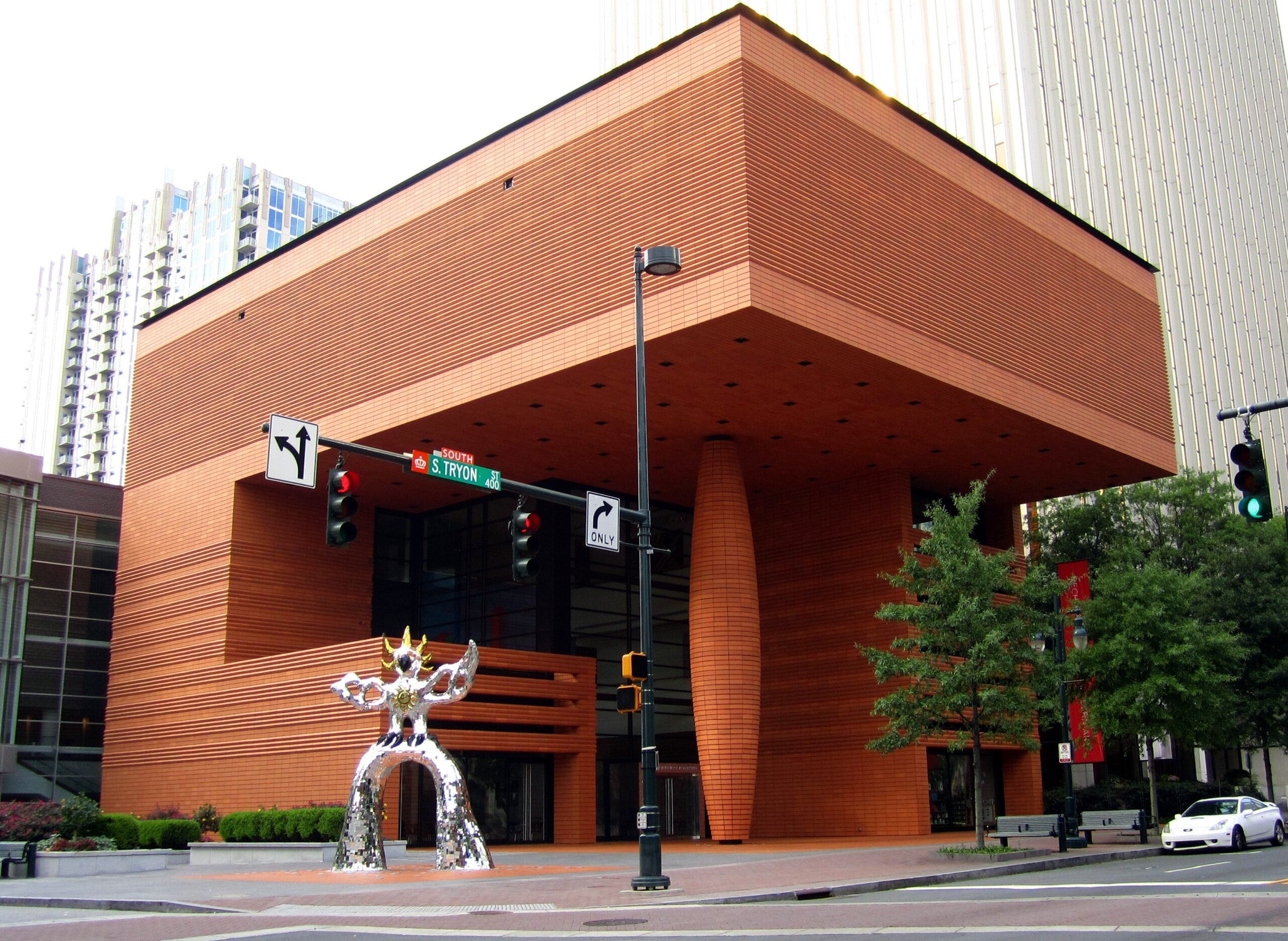 Orange brick modern building with layered facade and tall columns at a street intersection; a chrome sculpture in front and traffic signals nearby.