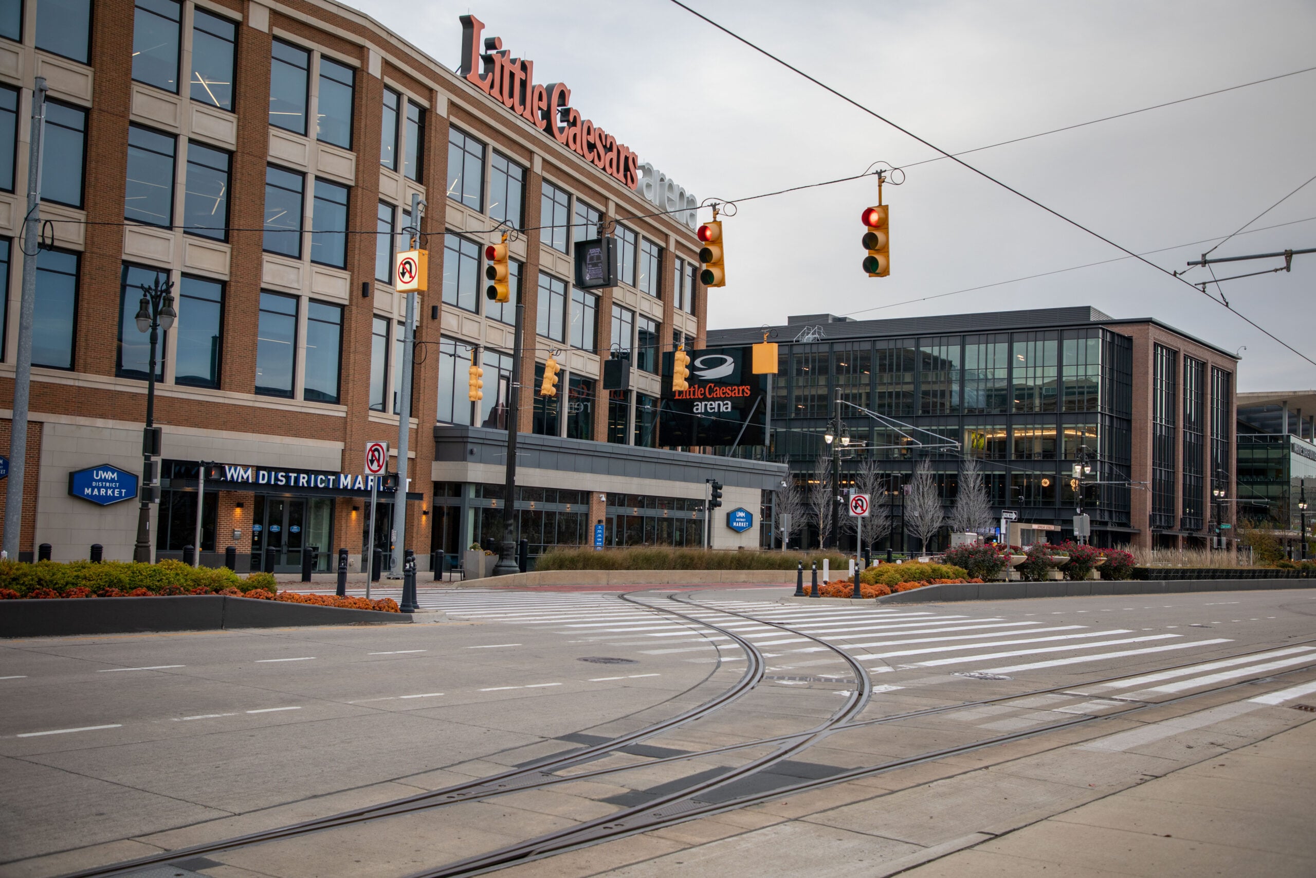 Street view of brick arena building with Little Caesars Arena sign, glass storefronts, and tram tracks in the road. downtown urban plaza feel with traffic lights overhead
