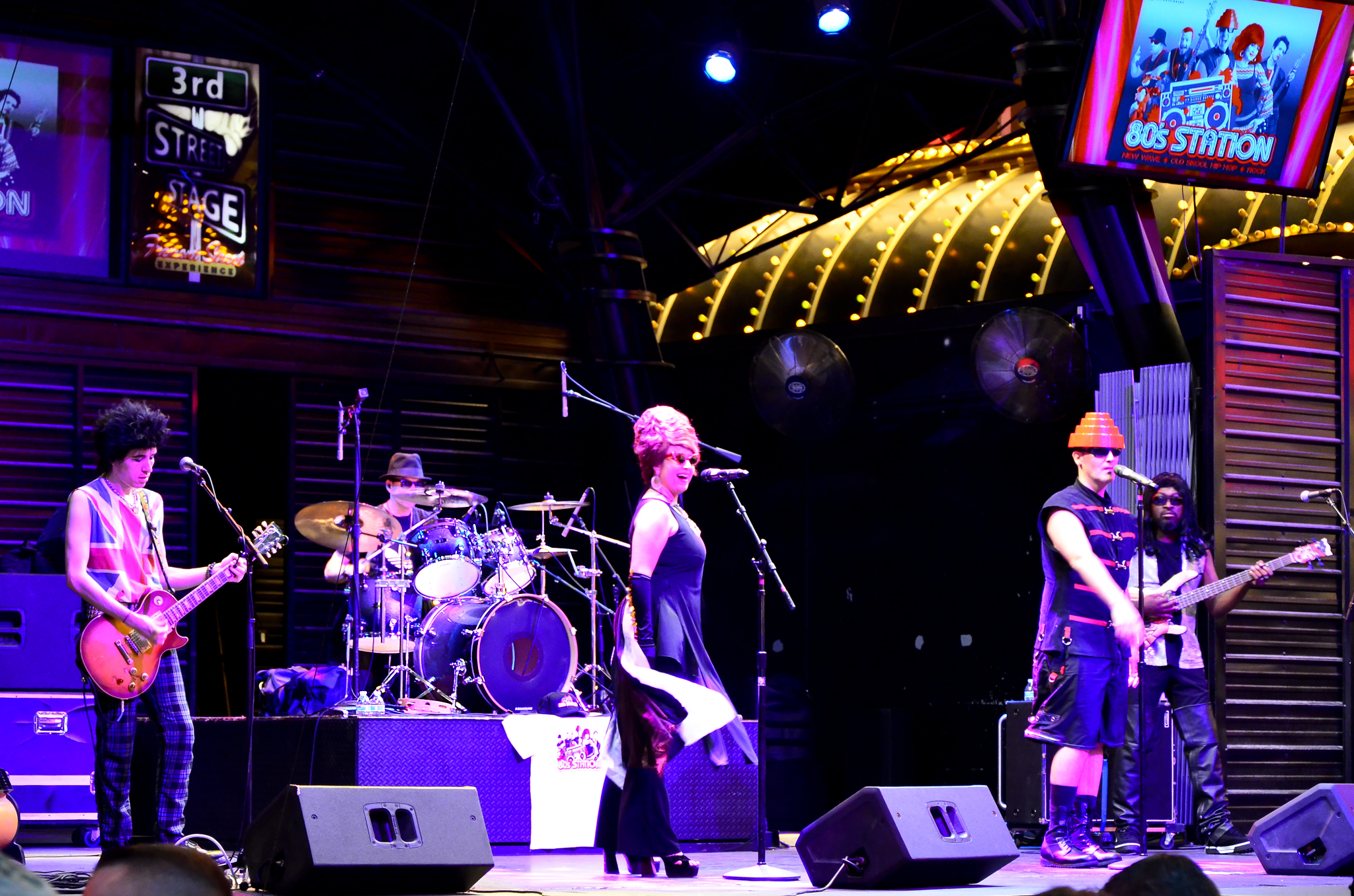 A live band performs on a neon-lit stage: guitarist front left, singer center, drummer rear, with audience in front.