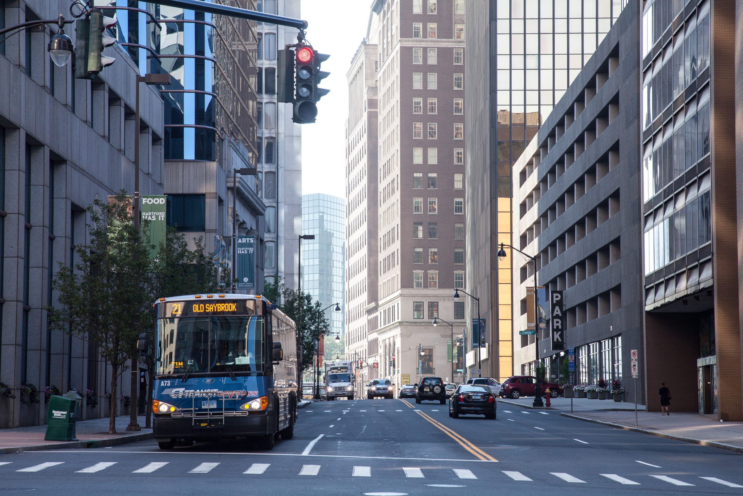 Blue city bus approaches at a busy urban street with tall office buildings on both sides and a crosswalk in the foreground.