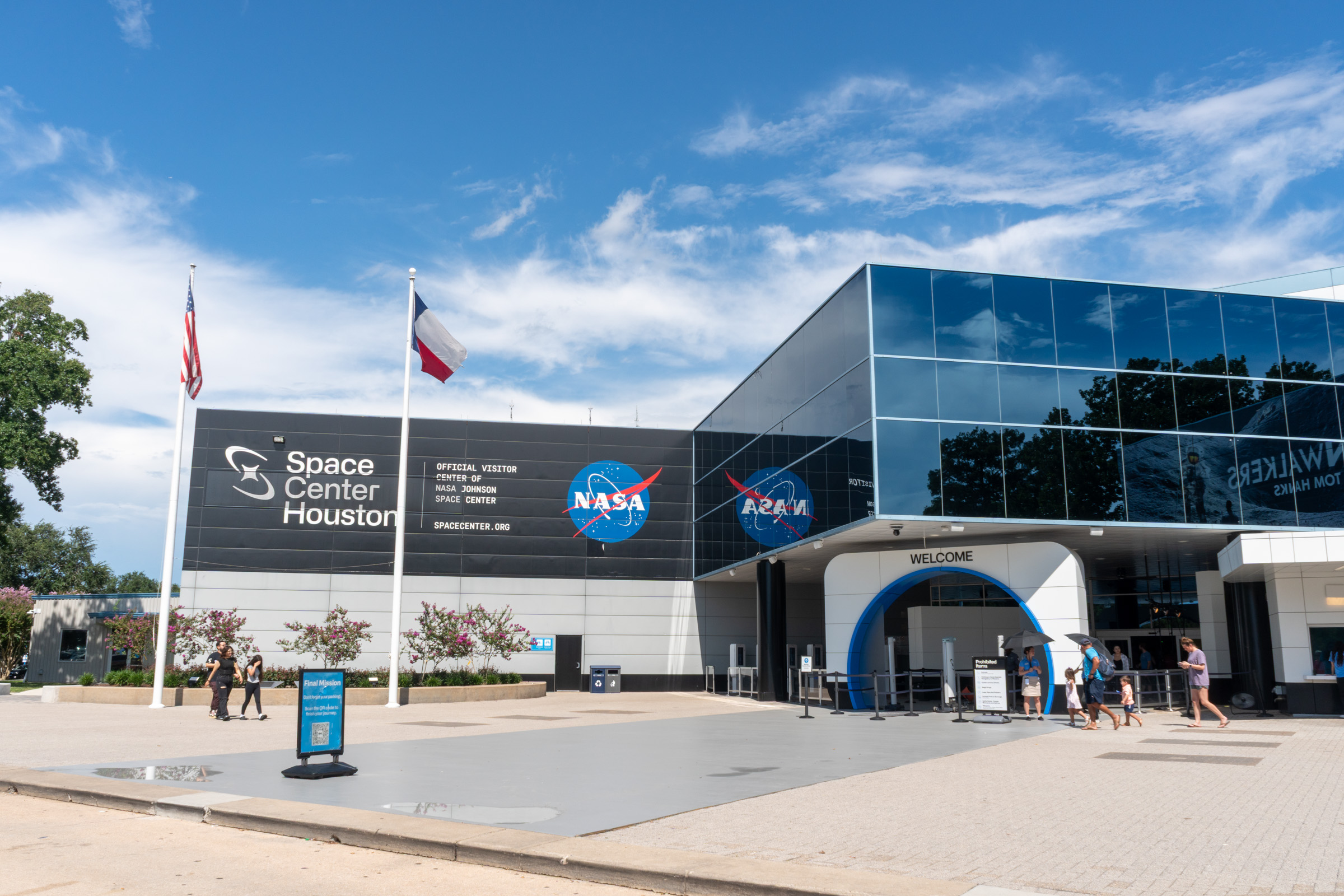- FIND-A-STAY Exterior view of Space Center Houston with NASA logos, American and Texas flags, and a glass-fronted building on a sunny day.