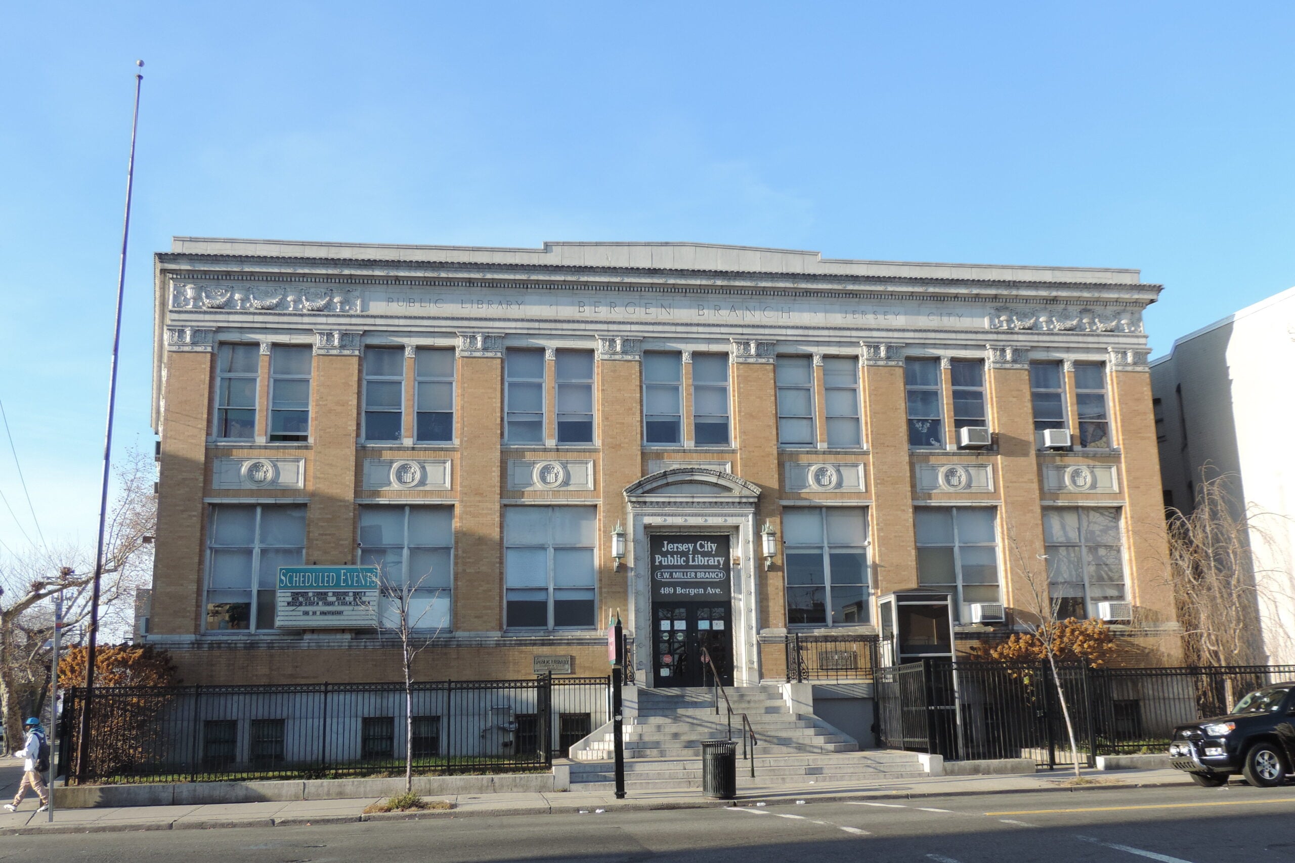 jersey-city-edmund-miller-library-bergen-avenue - FIND-A-STAY Front view of the Jersey City Public Library, Bergen Branch, a three-story brick building with decorative stone trim and a stairway entrance under a clear blue sky.