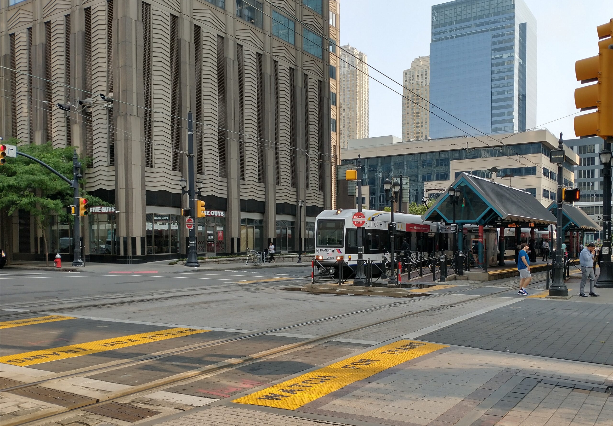 jersey-city-exchange-place-light-rail-july-2023 - FIND-A-STAY Urban city street with a white tram on tracks, pedestrians nearby, and tall modern buildings in the background.