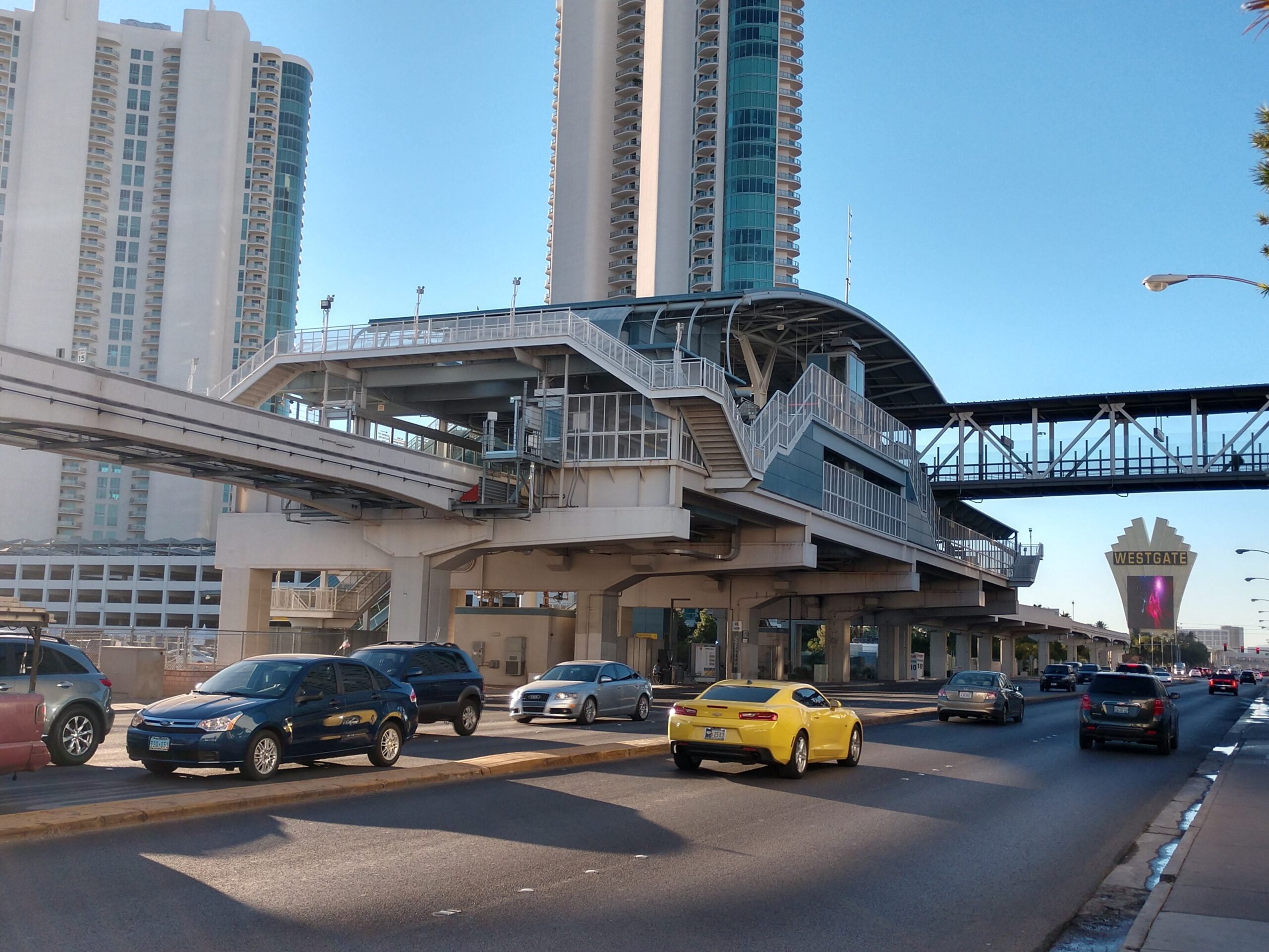 Urban scene with elevated pedestrian walkway over a multi-level roadway and tall white/blue glass high-rise buildings against a blue sky.",