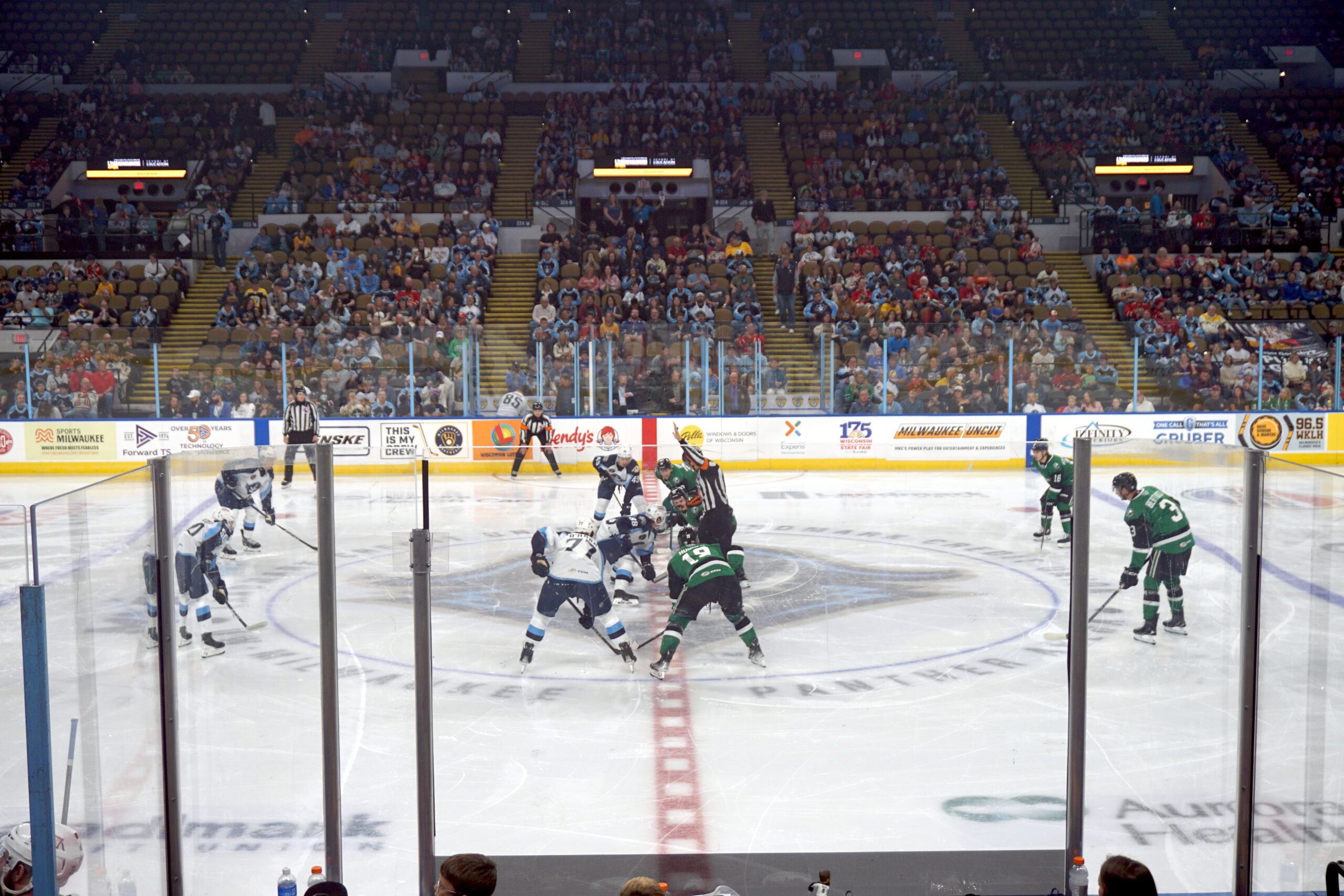 Ice hockey game underway with players contesting the puck near center ice, surrounded by a cheering crowd in the stands.
