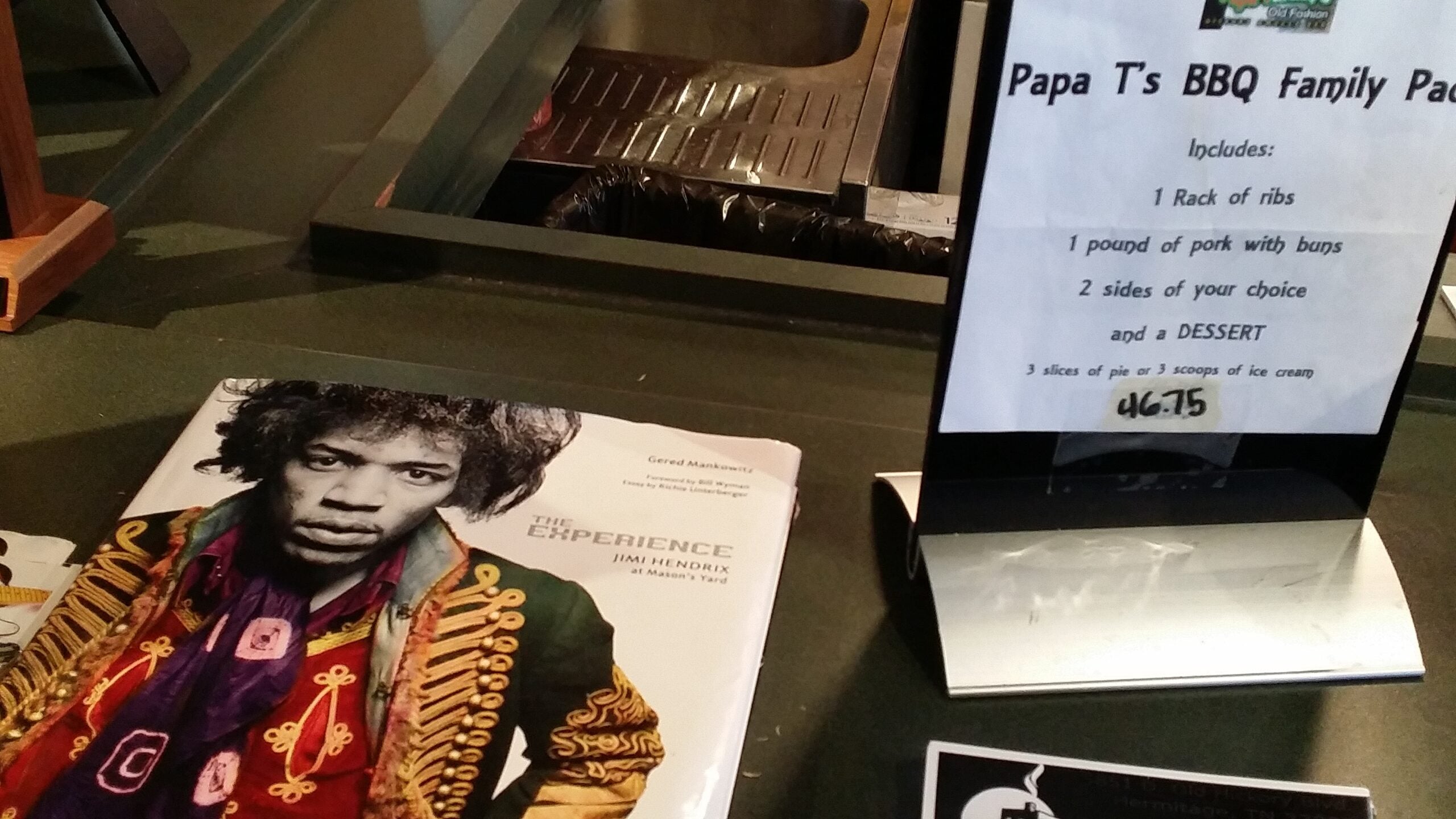 Display of books on a counter, including a Jimi Hendrix biography with a colorful cover, and a nearby sign advertising Papa T's BBQ Family Pack.