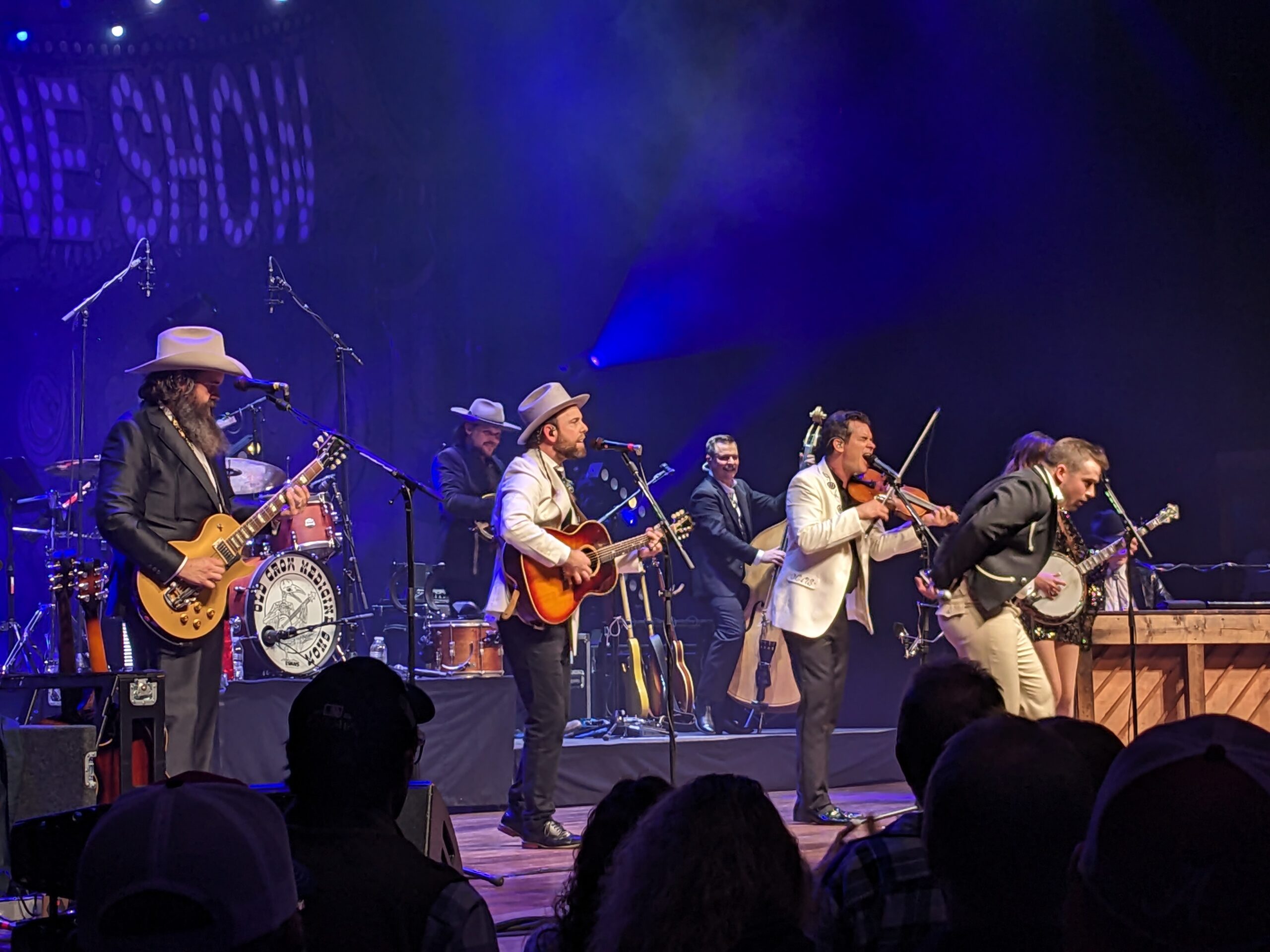 A group of musicians performing on a lit stage, many wearing suits and hats, playing guitars and violins while an audience watches from the foreground.