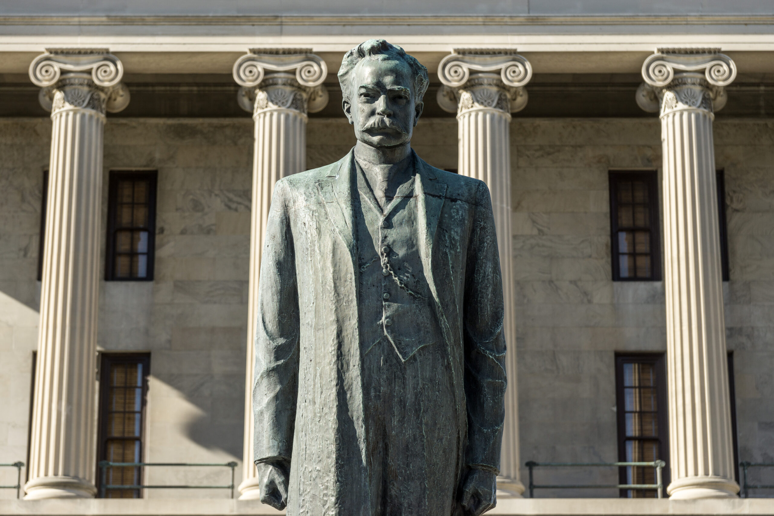 Bronze statue of a man in a suit standing before a neoclassical building with tall, decorative columns.