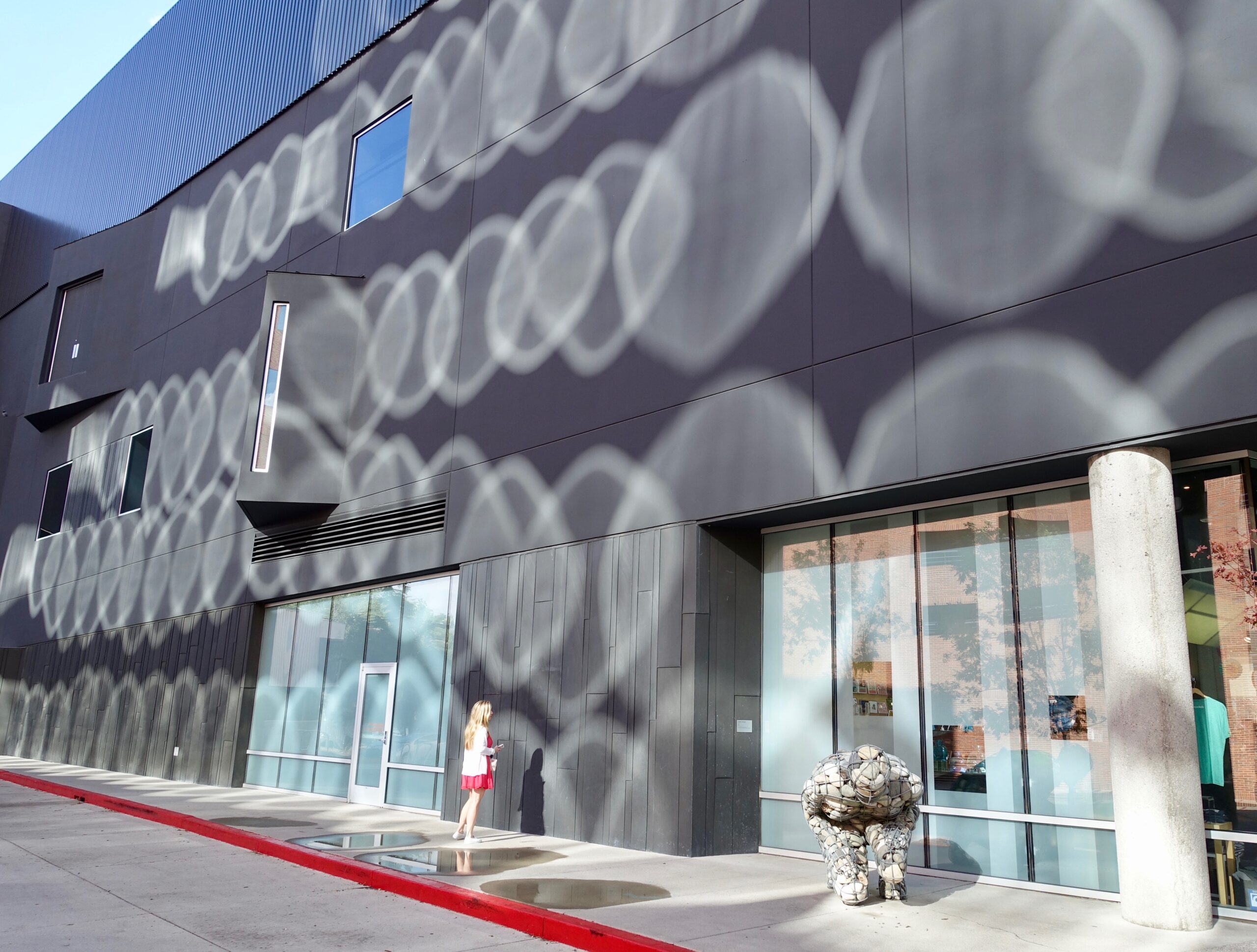 Modern grey building facade with circular reflective light patterns projected on panels; a woman in a red skirt stands near the entrance.