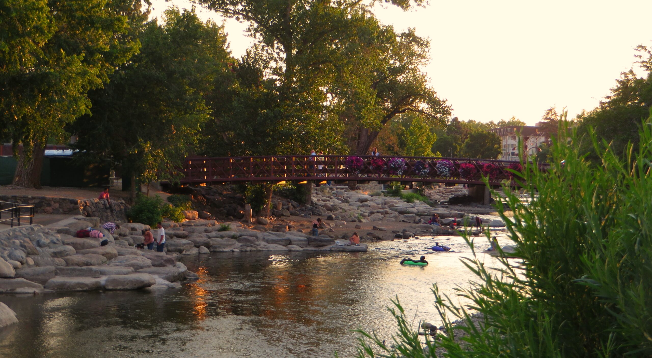 A wooden pedestrian bridge over a rocky riverbank, lined with colorful flower baskets and trees in a sunny park near the water.