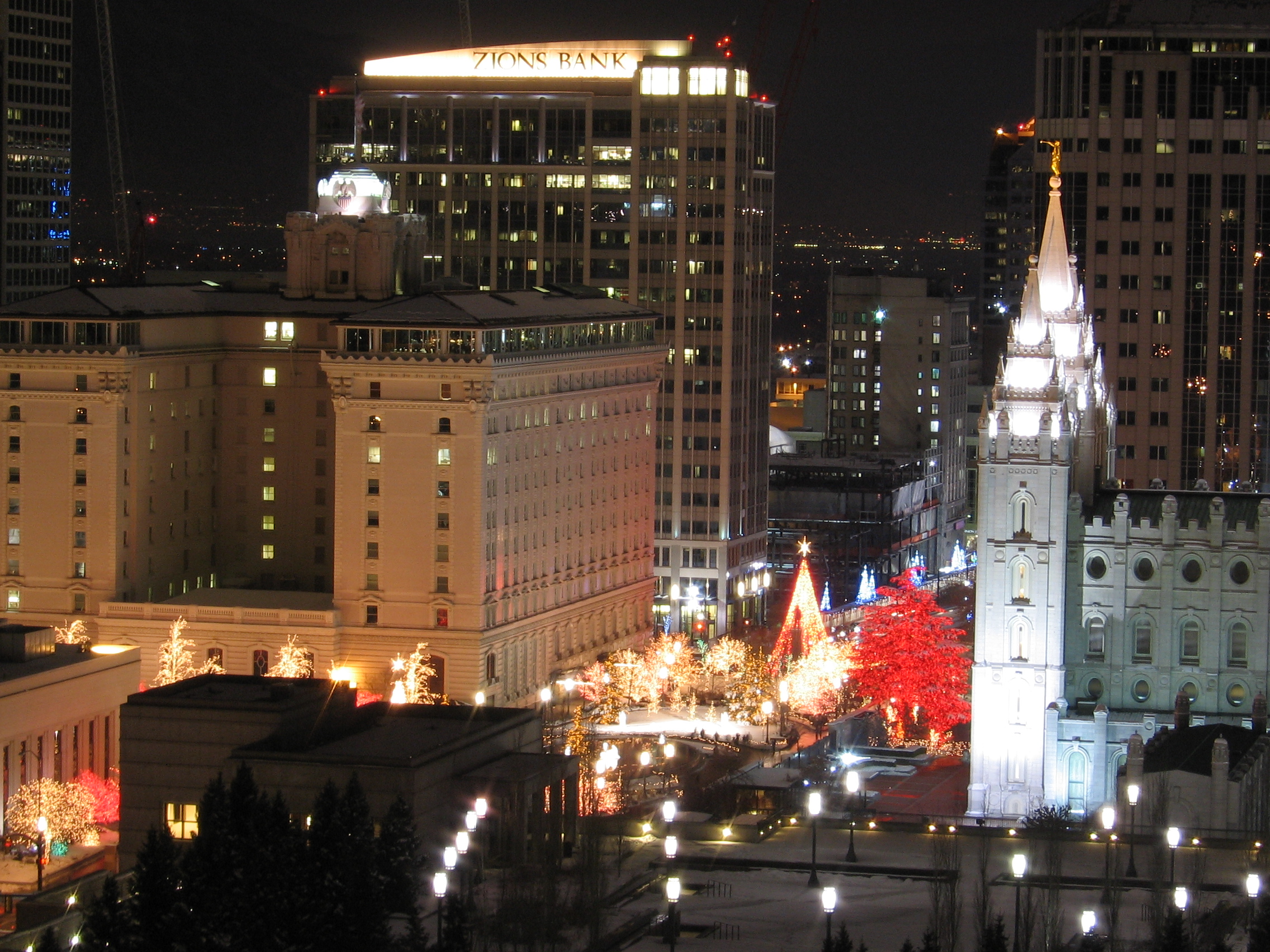 - FIND-A-STAY Nighttime downtown scene with festive Christmas lights along a plaza, a white historic building with a tall spire on the right, and modern office towers behind it.