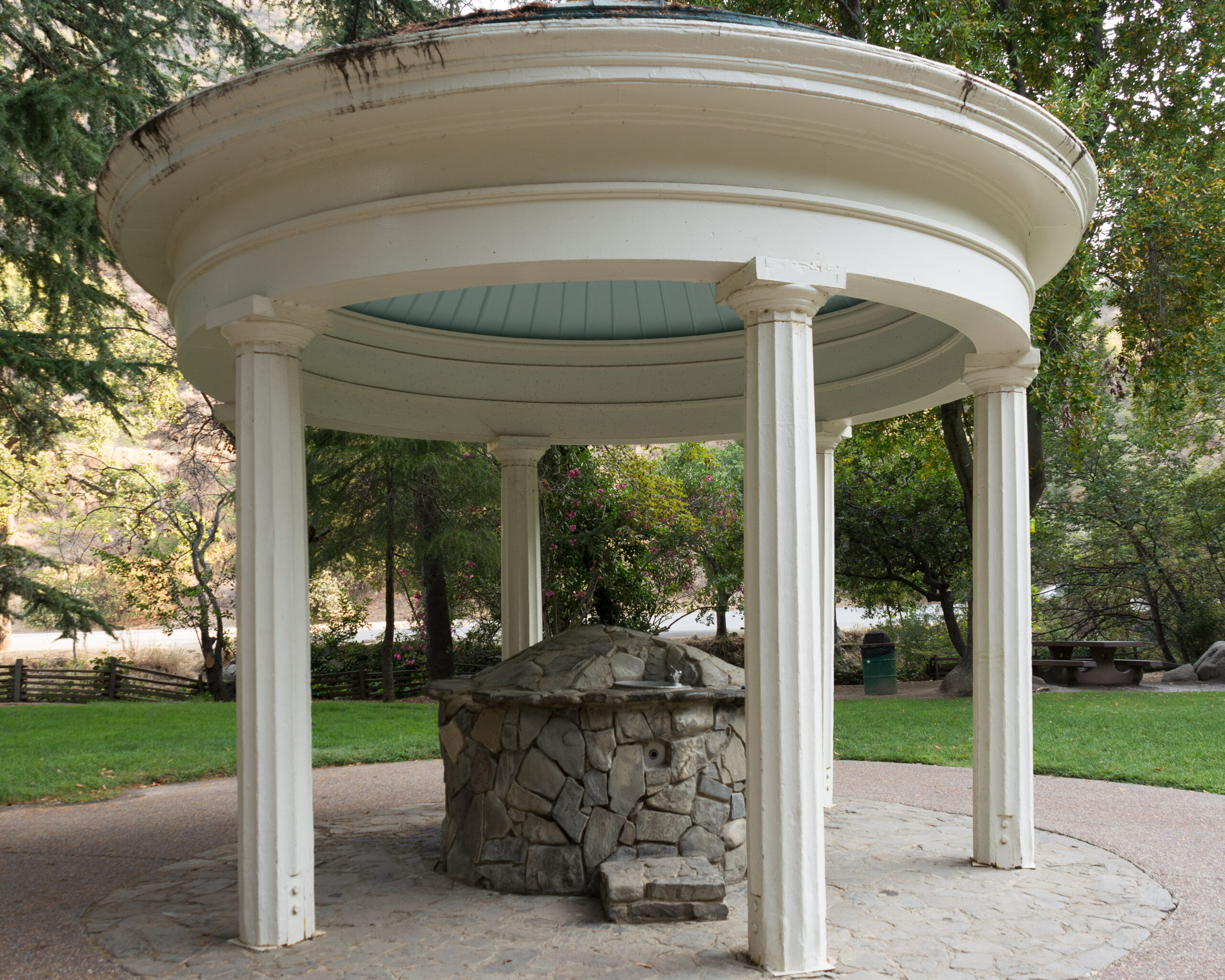 White circular gazebo with a stone well at the center in a park, surrounded by trees.