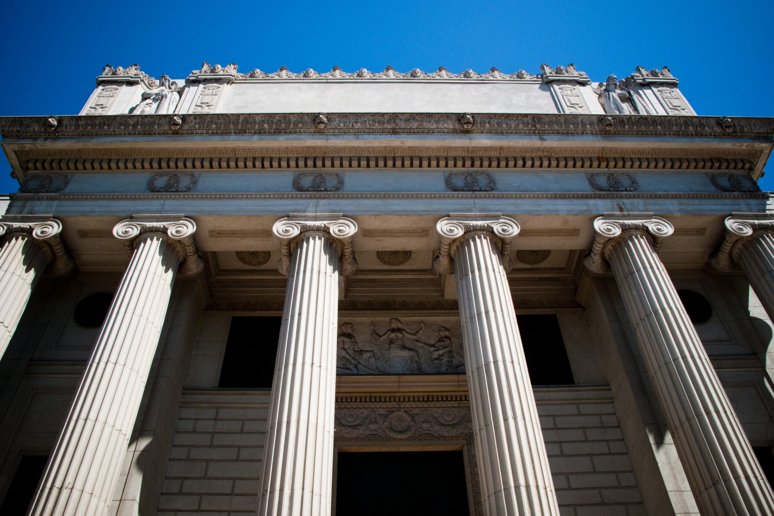 - FIND-A-STAY Front of a classical government building with tall fluted Ionic columns and decorative frieze under a clear blue sky.