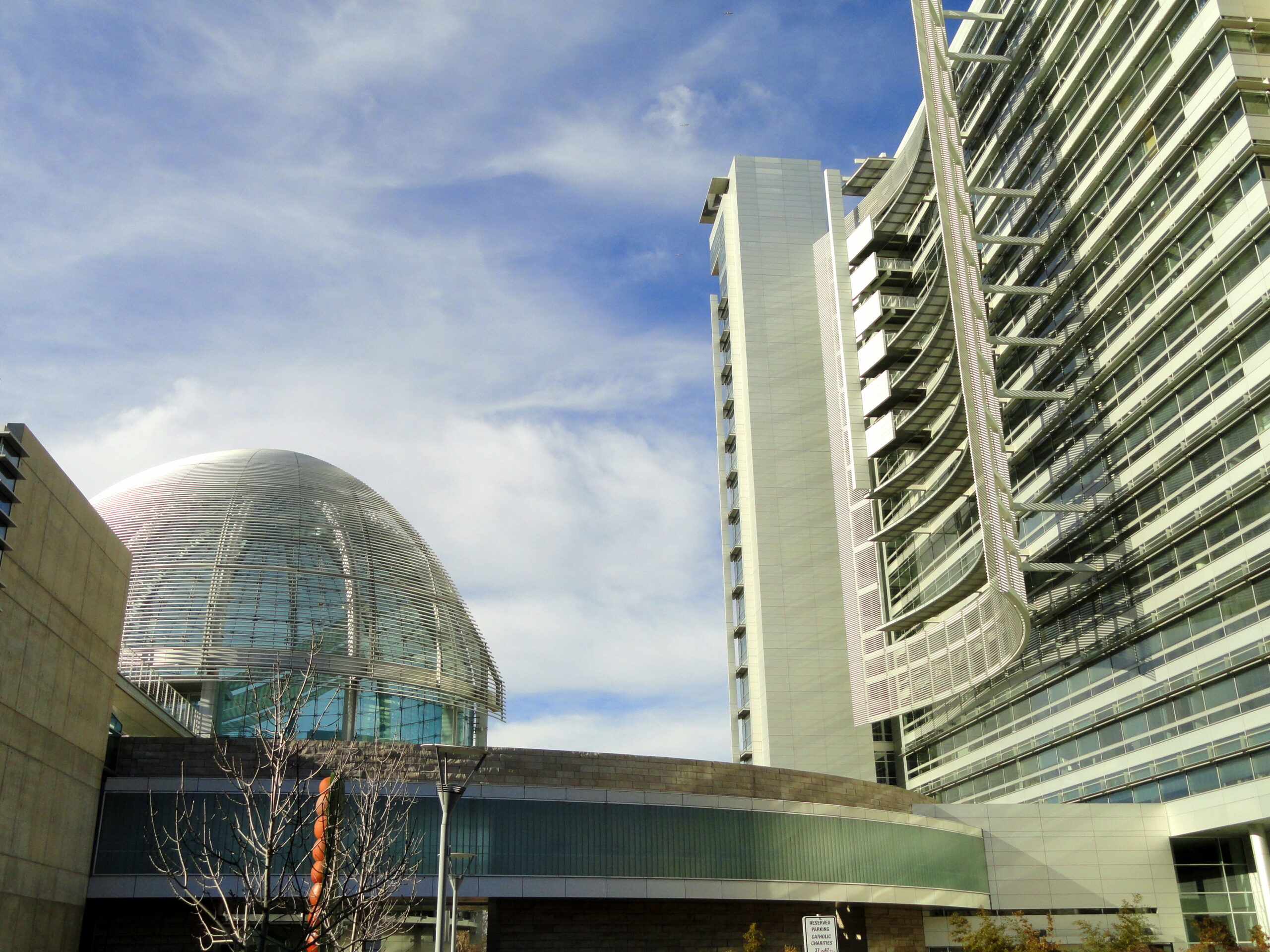 Modern architectural scene showing a large glass-domed sphere on the left and a tall glass-and-steel building with curved balconies on the right against a blue sky. (Informative)