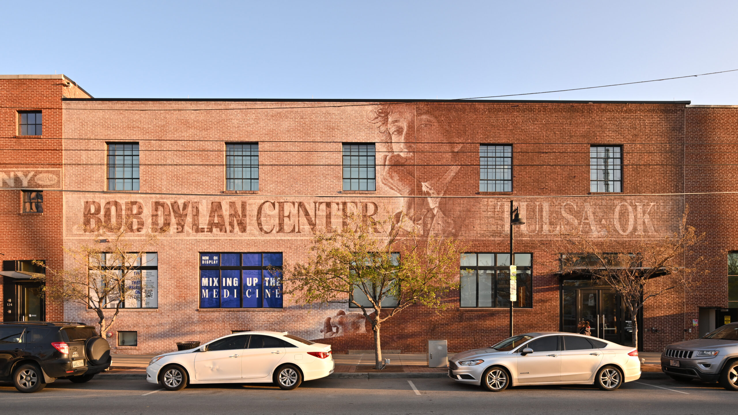 - FIND-A-STAY Brick building with a faded Bob Dylan Center mural across the facade and blue sky above; cars parked along the street in front.