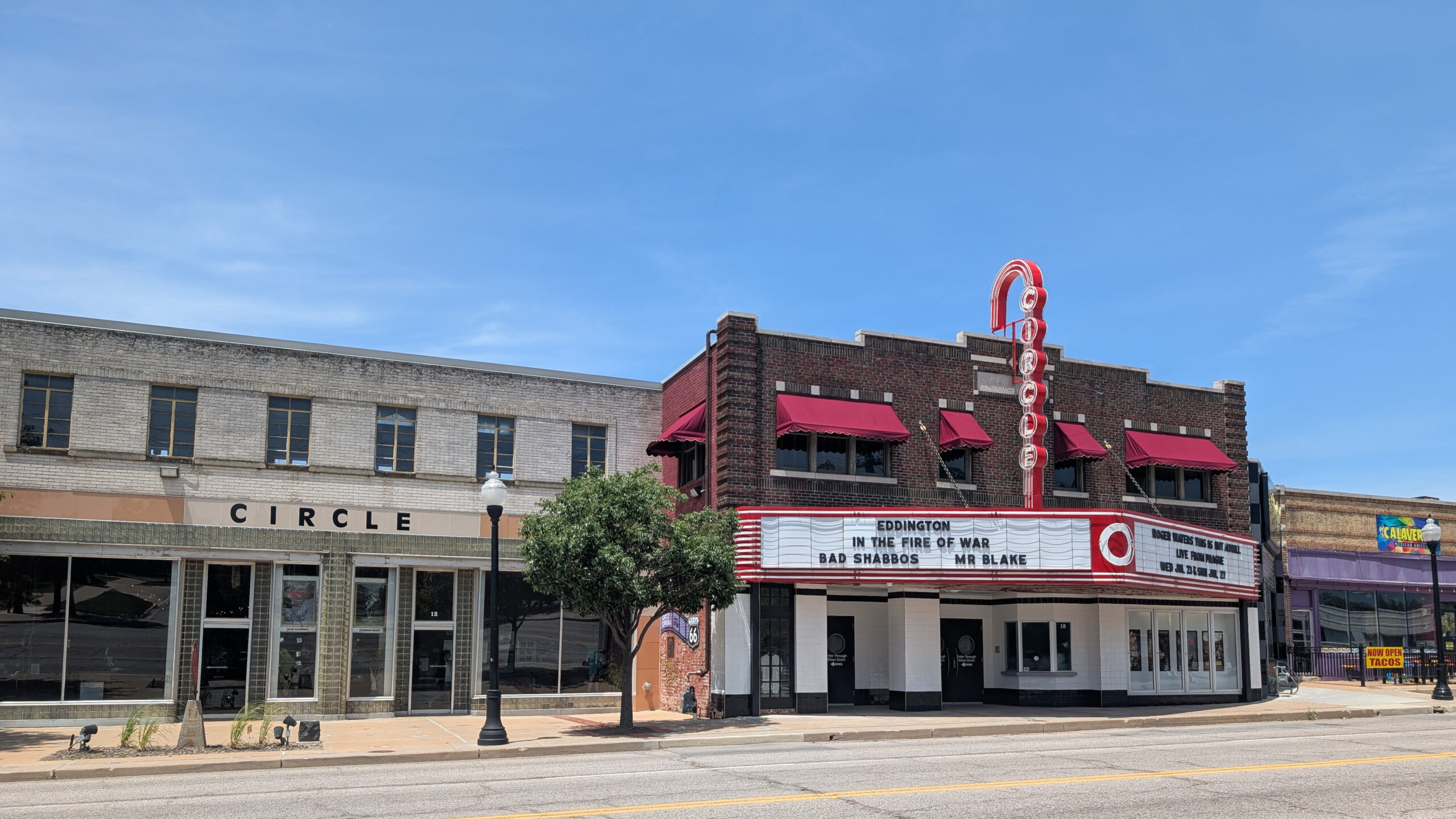Street view of a small downtown with a theater marquee, brick façade, red neon vertical sign, and red awnings on a sunny day