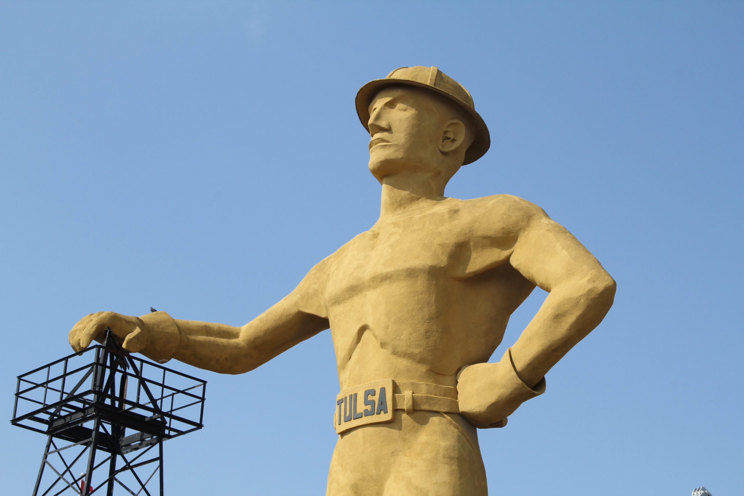 Golden statue of a man in a hat with arms outstretched against a clear blue sky, belt reads 'TULSA'.